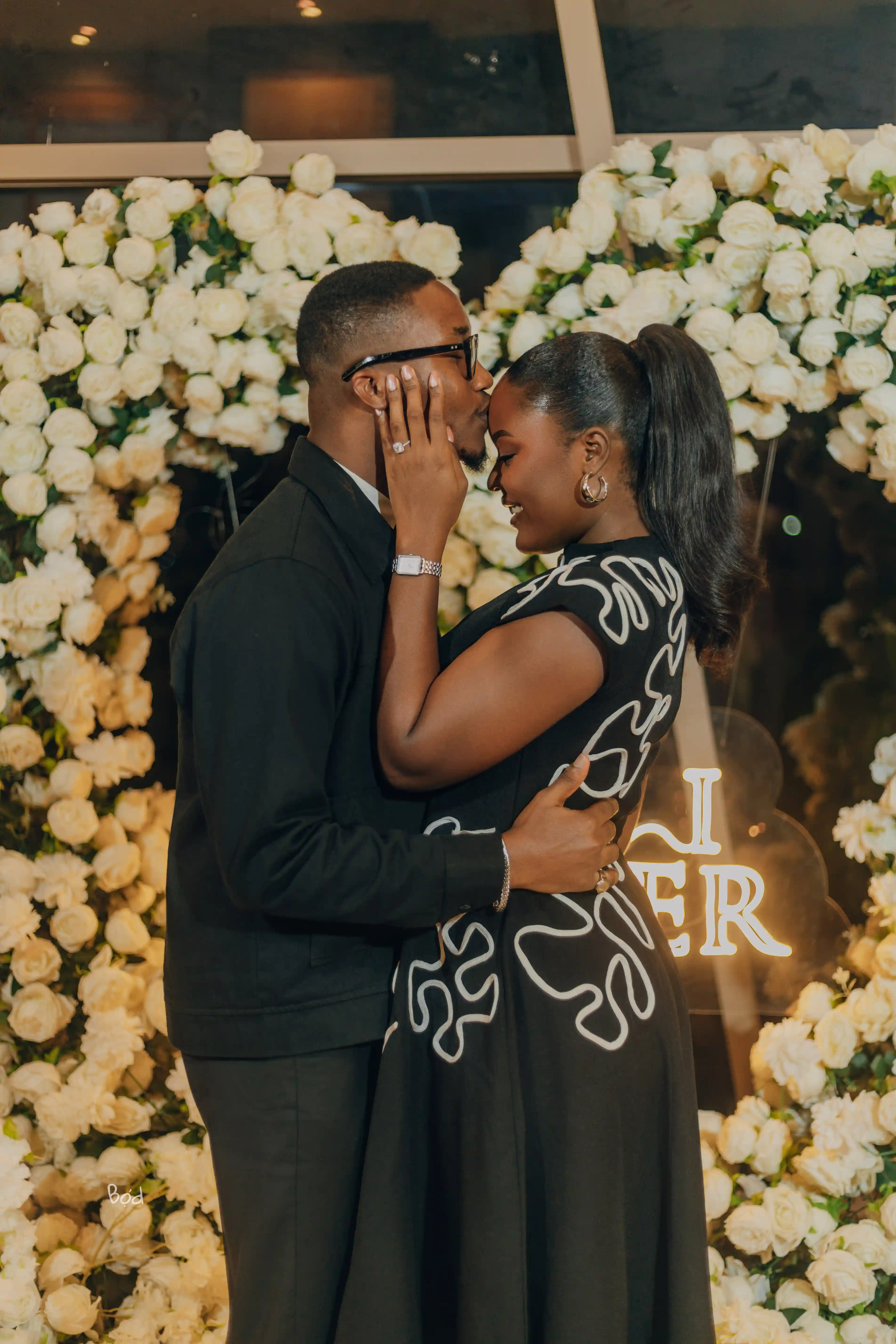 Kelvin kissing Priscilla's forehead in front of a white floral wall